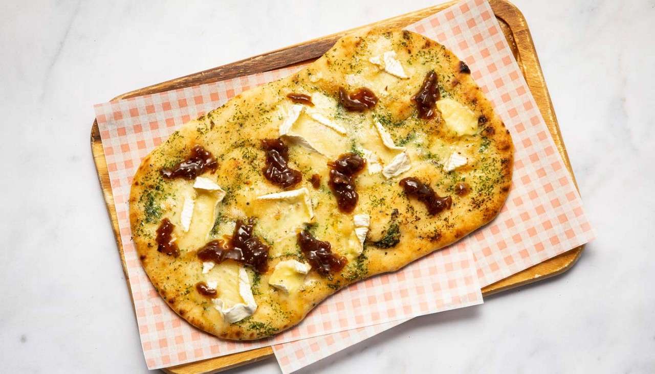 Flatbread rests topped with melted brie chunks, herb oil, and caramelized onion dollops, served on a wooden board lined with pink-check paper, set on a white marble surface.