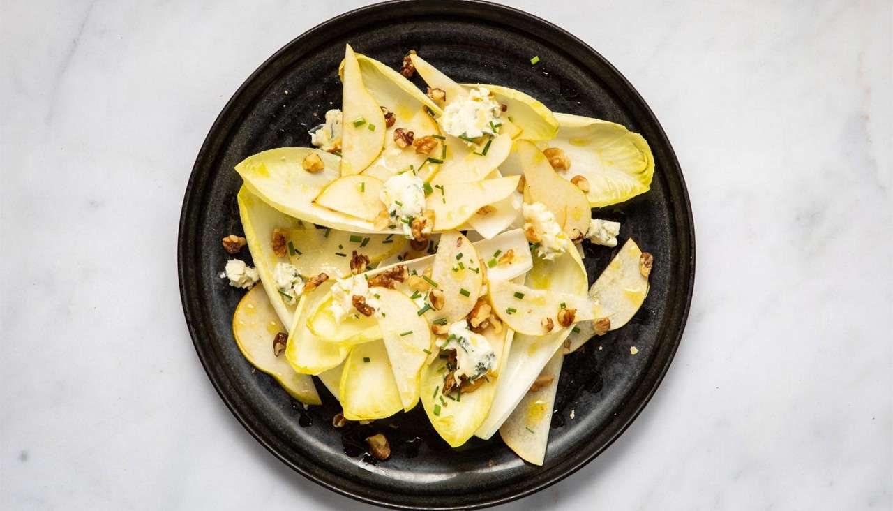 Endive and pear salad rests on a black plate, topped with blue cheese crumbles, walnuts, chopped chives, and olive oil, sitting on a white marble surface.