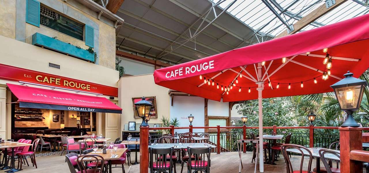 Red café umbrellas shade bistro tables while string lights hang, in an indoor atrium outside a restaurant facade. Visible text: CAFE ROUGE; OPEN ALL DAY; CAFE • BISTRO • RESTAURANT; Est. 1989; TOILETS.
