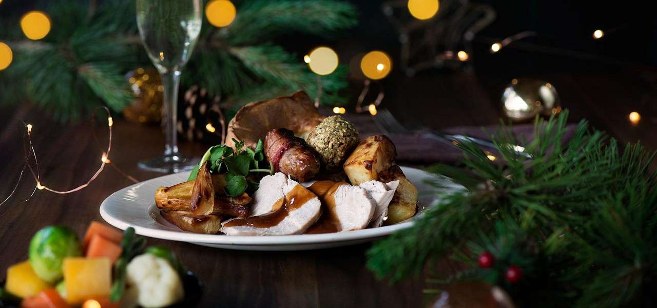 Turkey slices with gravy rest on a plate, accompanied by roast potatoes, stuffing, and bacon-wrapped sausages, on a festive table with pine branches, fairy lights, and a champagne flute.