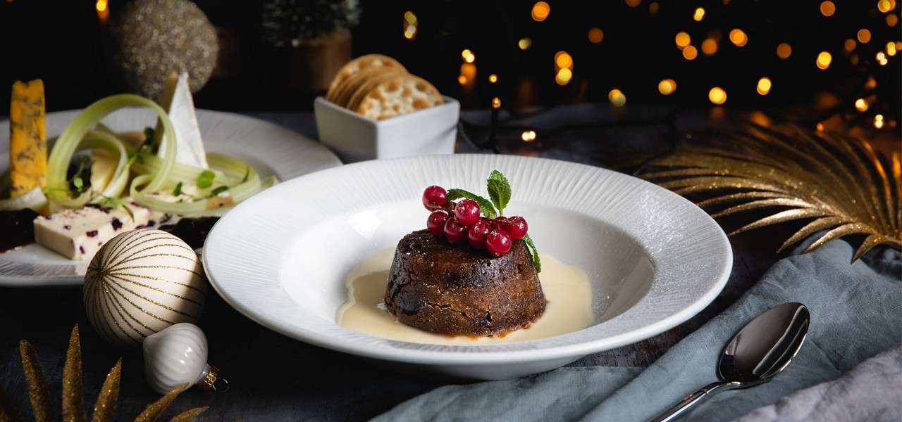 Plated dessert sits in custard on a white bowl, garnished with red currants and mint; nearby cheese, crackers, ornaments, and a spoon; warm festive lights glow in the dark background.