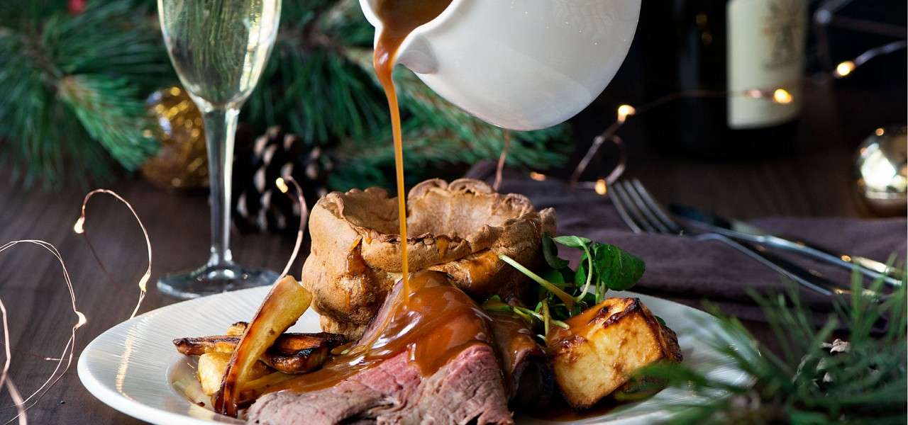 Roast dinner plate receives gravy poured from a white jug onto sliced beef. Yorkshire pudding, roast potatoes, parsnips, greens rest on a festive table with champagne flute, fairy lights.