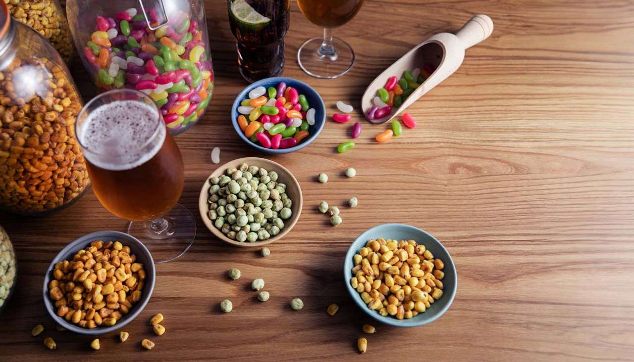 Assorted snacks rest on a wooden table: bowls of roasted corn and wasabi peas, jelly beans spilling from a scoop, large jars of treats, and glasses of beer and cola.