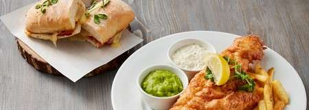 Fried fish and chips sit on a white plate with tartar sauce and mushy peas; nearby, a toasted ciabatta sandwich rests halved on a wooden board atop a gray tabletop.