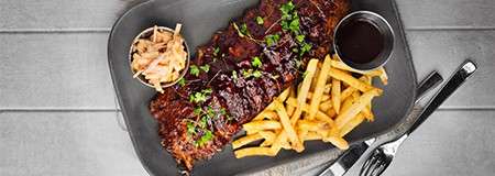 Barbecued ribs sit glazed with sauce, garnished with herbs, accompanied by fries, coleslaw cup, and dipping sauce on a dark plate, placed on a gray table beside fork and knife.