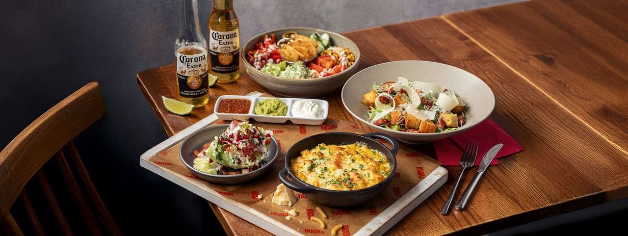Steak rests topped with melting herb butter, accompanied by cup of fries, grilled tomato halves, and green salad; served on a dark platter on a wooden table with side dishes.