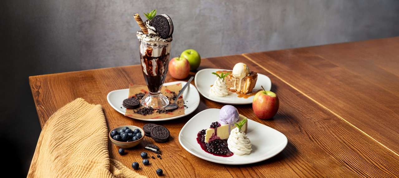 Milkshake overflows with whipped cream and chocolate drizzle, garnished with wafer sticks and a skewer of doughnut and strawberry, sitting on a white plate with chocolate syrup on wooden table.