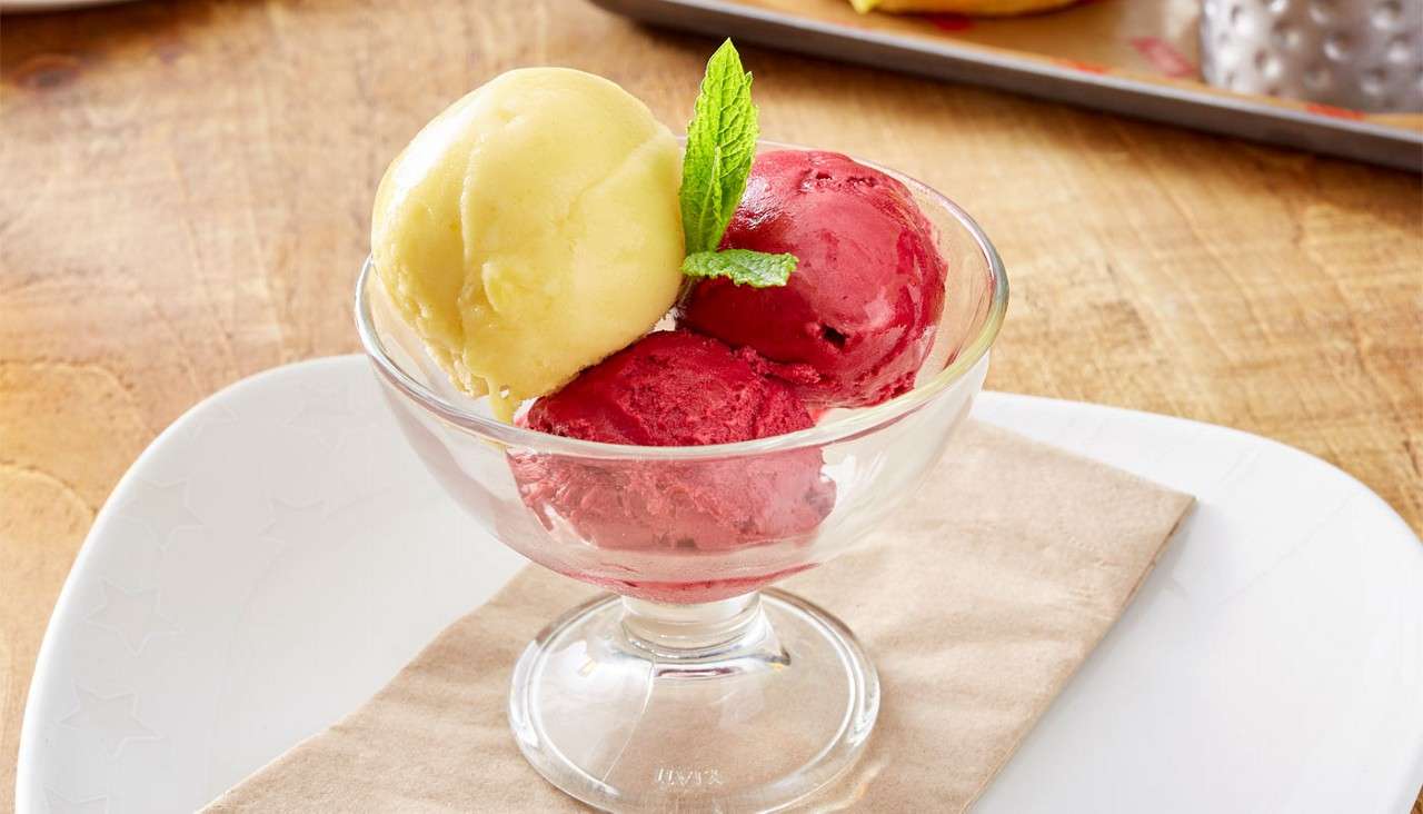 Scoops of sorbet sit in a glass dish, garnished with a mint sprig; one pale yellow and two red; placed on a napkin-lined plate on a wooden table.