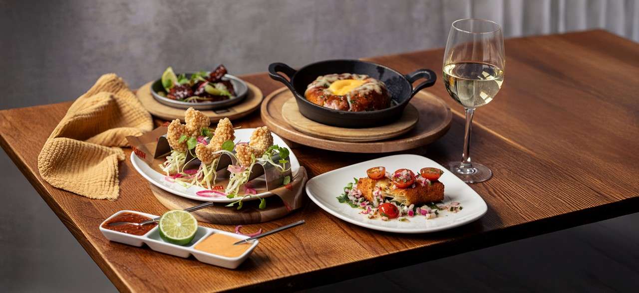 Salad bowl rests on a table, displaying leafy greens, corn, red onion, and tomato pieces, surrounded by other side dishes like fries and another salad in a restaurant-style setting.
