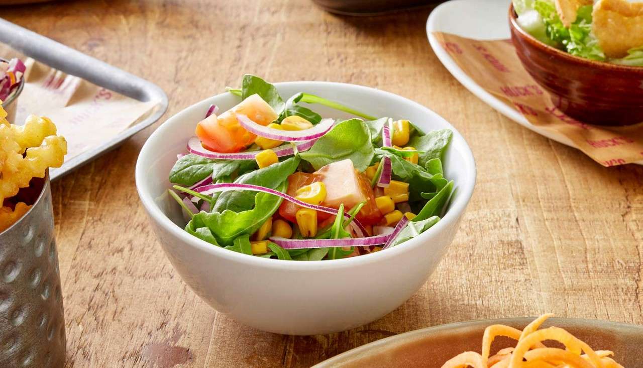 Salad bowl rests on a table, displaying leafy greens, corn, red onion, and tomato pieces, surrounded by other side dishes like fries and another salad in a restaurant-style setting.