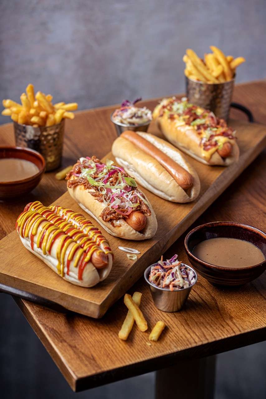 Bowl of nachos holds salsa, shredded cheese, guacamole, jalapeños, and chips, surrounded by ramekins of green herby mixture, salsa, sour cream, olives, and more chips on a wooden table; tray paper reads: “HUCK’S”.