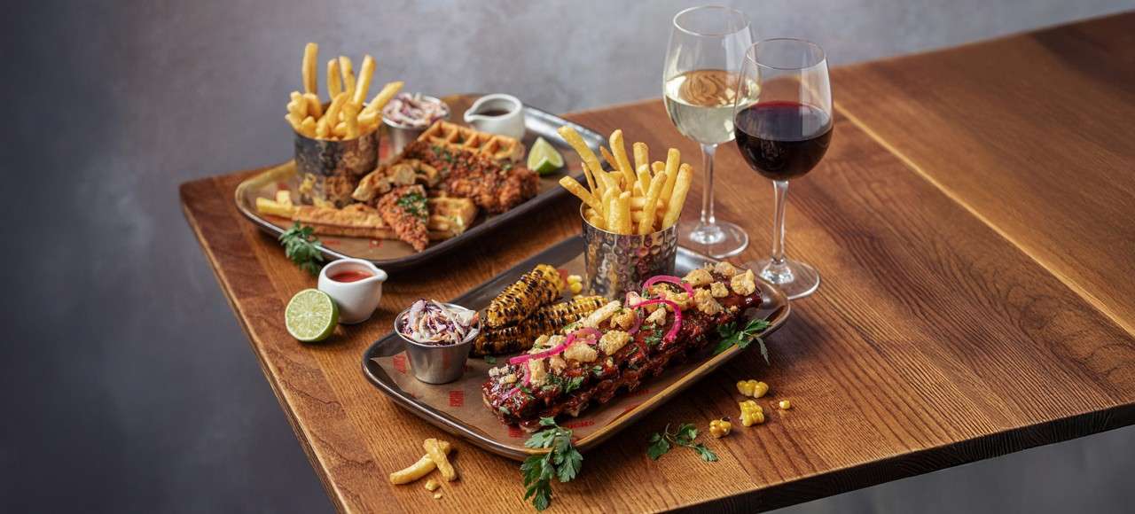 Rack of barbecue ribs sits on a platter, garnished with microgreens, with fries, coleslaw, and dipping sauce; nearby, a beer glass and cutlery rest on a wooden table.