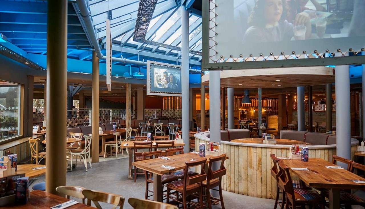 Restaurant dining area—empty tables set with condiments—sits beneath a glass skylight and blue beams, with wooden booths, mixed chairs, and hanging screens showing images in a spacious, warmly lit interior.