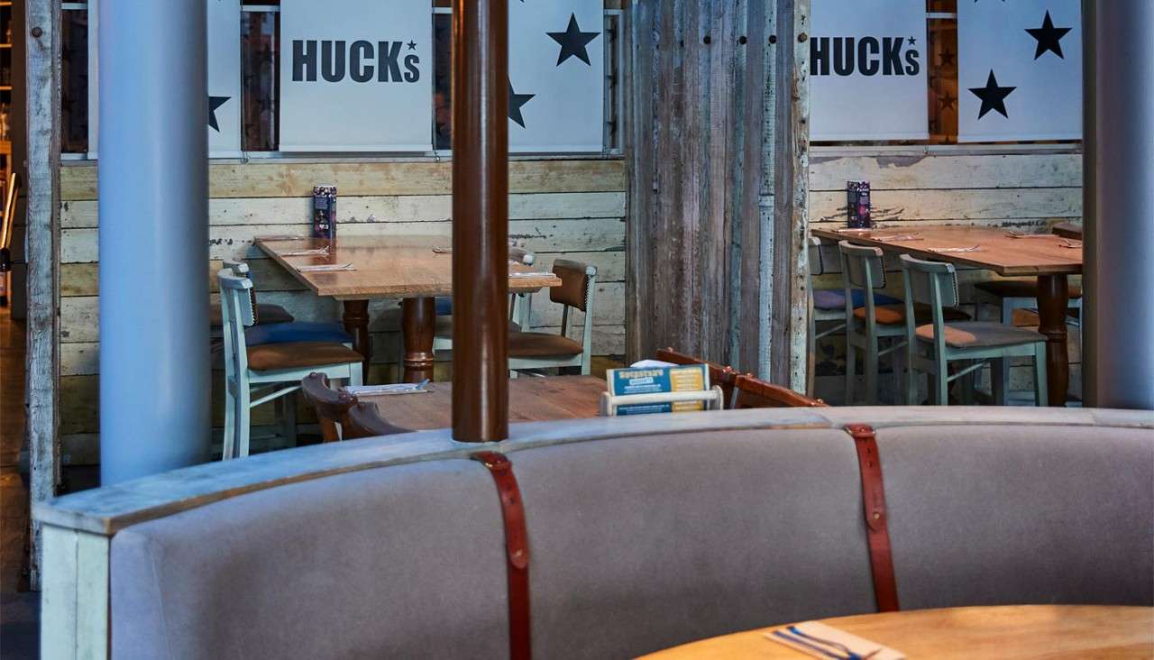 Curved booth seats encircle a wooden table, holding menus; nearby wooden tables sit empty. Rustic restaurant setting with slatted walls and partitions reading: HUCK's, HUCK's.