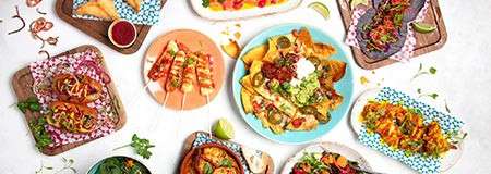 Assorted Mexican dishes sit arranged on a table; tacos, nachos with guacamole and sour cream, skewers, grilled corn, and lime wedges surround colorful plates, suggesting a festive shared meal.