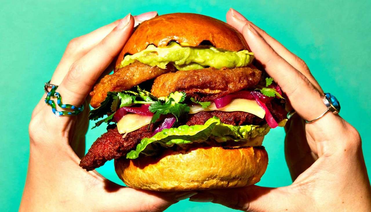 Burger stacked with lettuce, cheese, pickled onions, cilantro, fried onion rings, guacamole, and a patty, being held with both hands against a turquoise background, inside a glossy brioche bun.