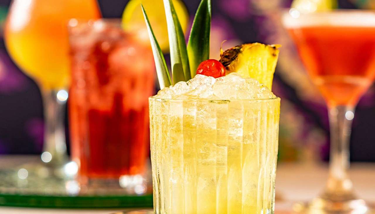 Yellow tropical cocktail sits on crushed ice, garnished with pineapple wedge, cherry, and leaves; blurred colorful cocktails surround it on a bar table with warm lighting.