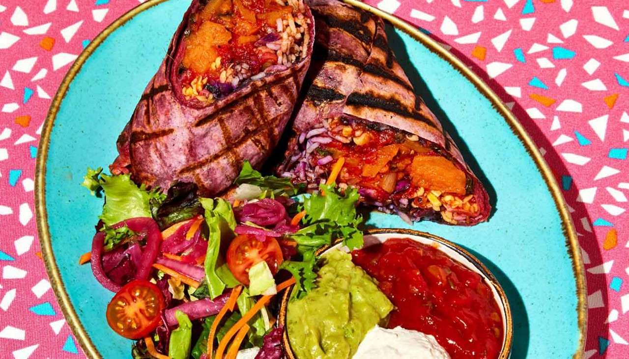 Grilled burrito halves sit cut open, revealing rice, beans, vegetables, and sauce. They rest on a turquoise plate with mixed salad, guacamole, salsa, and sour cream atop a speckled tablecloth.