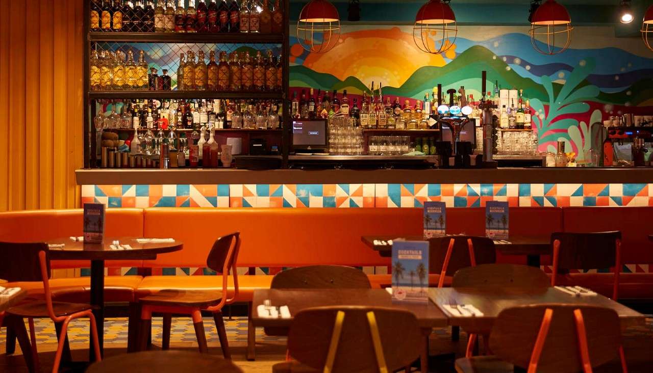 Bar counter displays liquor bottles, glasses, and taps on lit shelves; context: vibrant restaurant with orange banquettes, wooden chairs, geometric tiles, and a colorful mural of hills and sun.