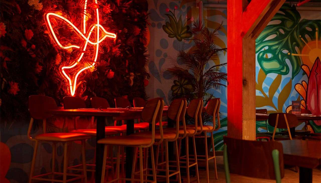 Neon bird sign glows, casting red light over high-top chairs and tables, against a leafy wall. Surrounding context: tropical mural, indoor plants, wooden beam, dimly lit restaurant-bar setting.