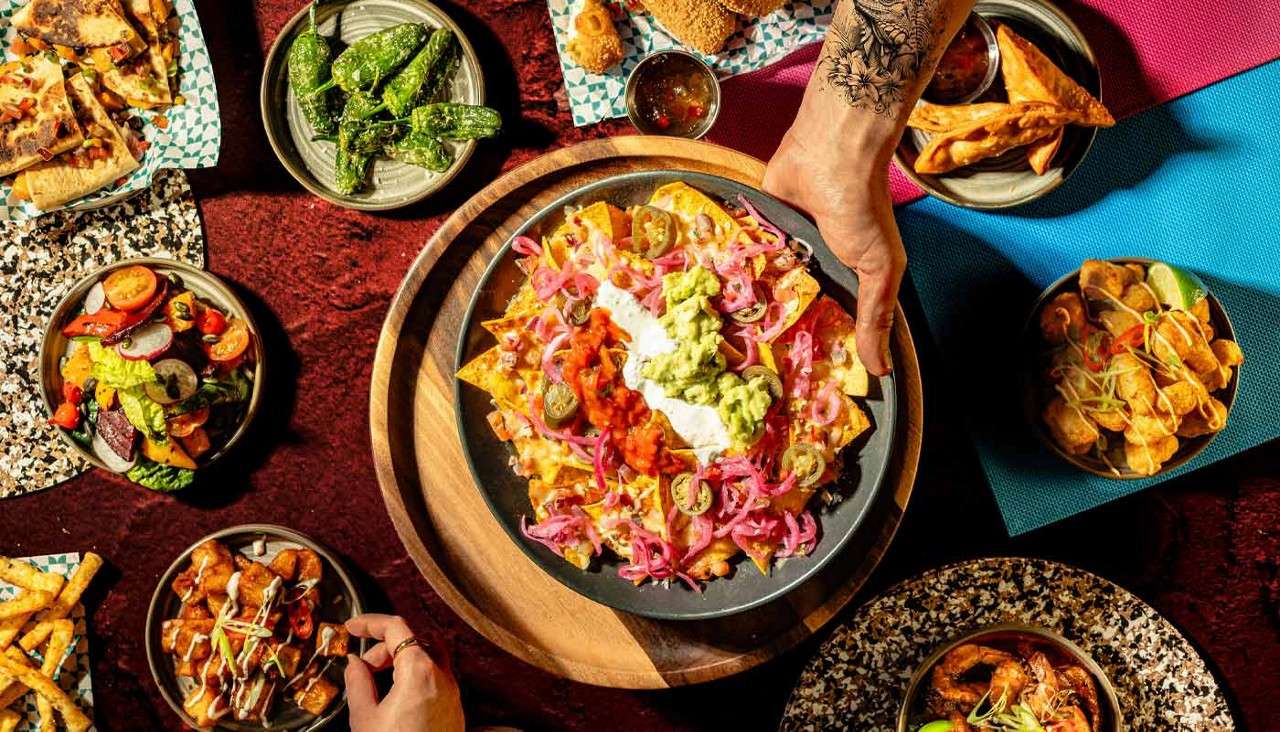 Nachos topped with guacamole, sour cream, salsa, pickled onions, and jalapeños, served by a tattooed hand, surrounded by assorted small plates—salad, peppers, fries, dumplings, and shrimp—on a colorful tabletop.