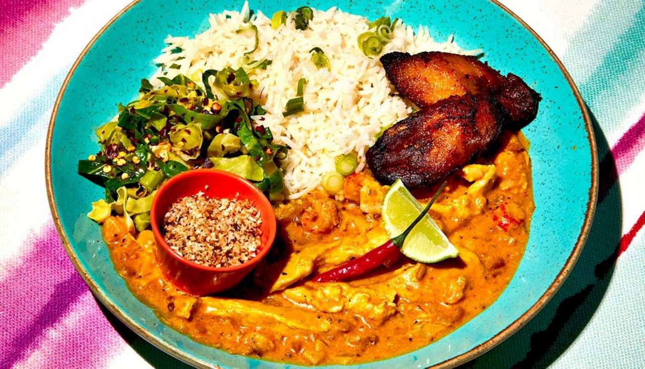 A turquoise plate presents chicken curry, rice with scallions, sautéed greens, fried plantains, a lime wedge, red chili, and a bowl of coconut seasoning, on a striped tablecloth.