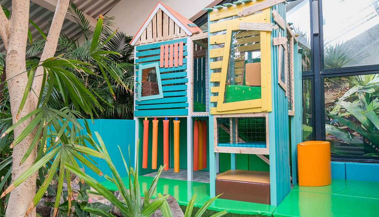 Colorful wooden playhouse stands with slanted windows, mesh panels, and hanging foam posts; children can climb and crawl. Surrounded by tropical plants and glass walls in an indoor recreational space.