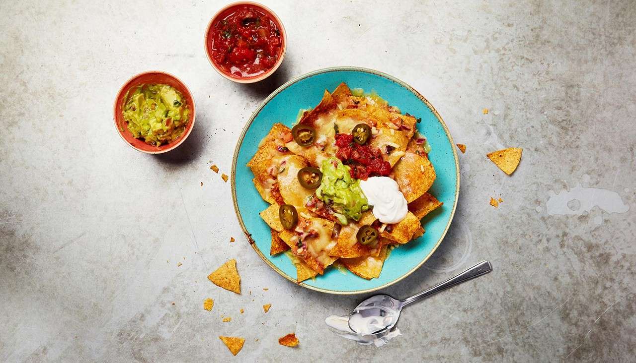 Nachos sit topped with melted cheese, jalapeños, salsa, guacamole, and sour cream, arranged in a turquoise plate; side bowls of guacamole and salsa on a gray tabletop with crumbs.