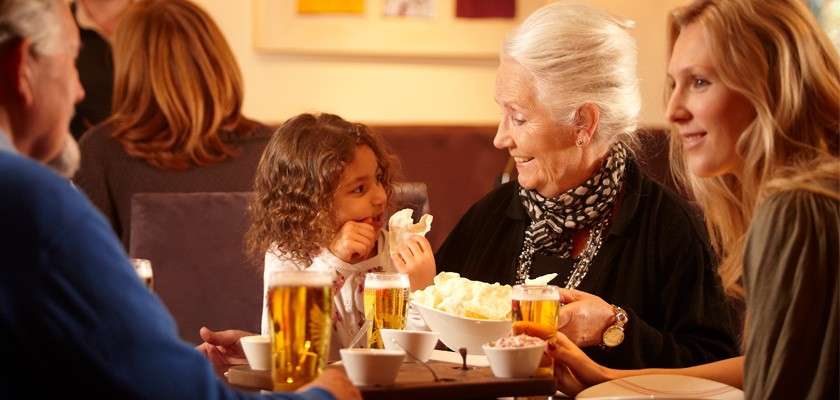 Older woman and young girl share snacks, smiling, at a restaurant table with beer glasses, bowls, and plates, while two adults chat nearby in a warmly lit indoor dining setting.