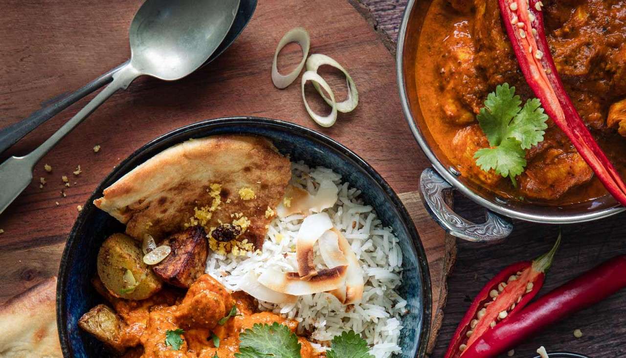 Bowl of curry and rice with naan and roasted potatoes, garnished with cilantro and coconut; nearby, metal pot of curry, red chilies, onion rings, and spoons on a wooden table.