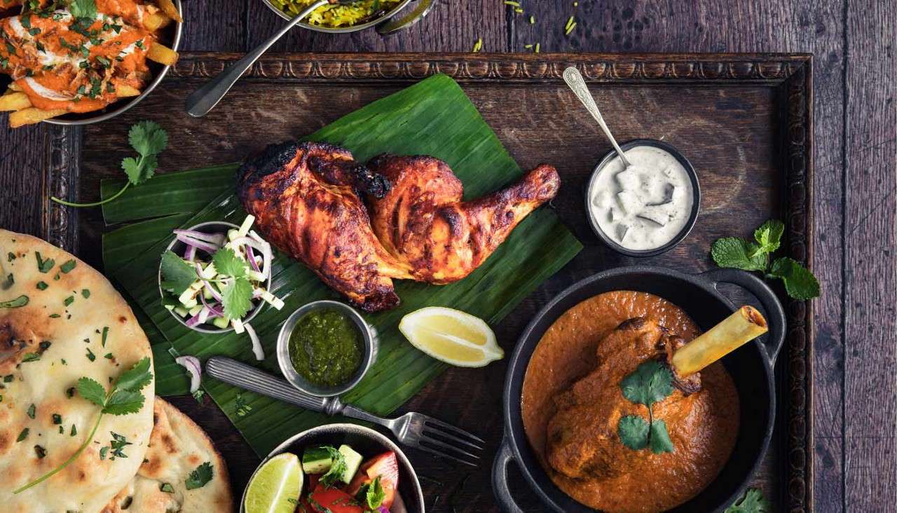 Grilled chicken rests on banana leaf, accompanied by curry lamb shank in pot, naan, rice, fries with sauce, salads, chutneys, lemon wedges; arranged on rustic wooden tray tabletop.