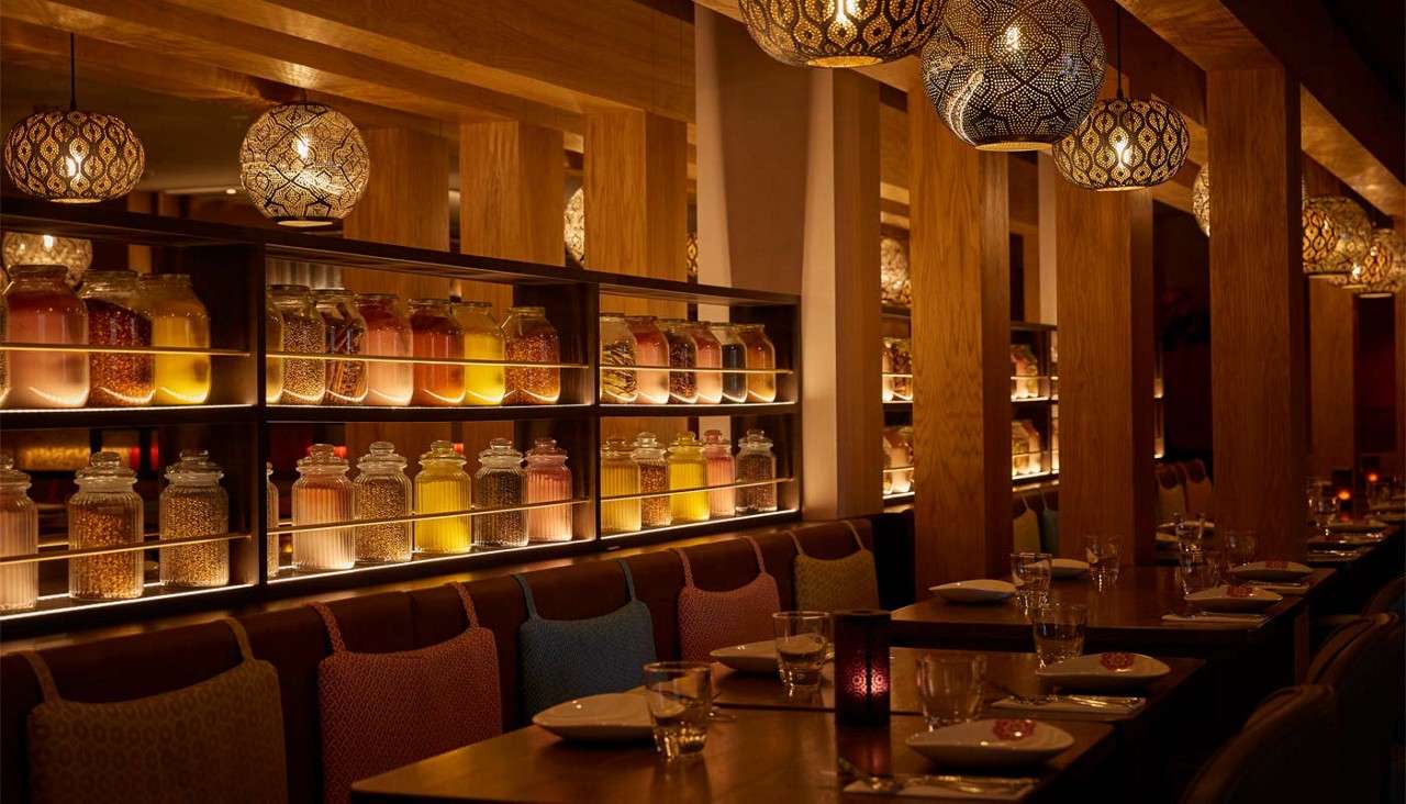 Backlit glass jars of colorful grains and liquids glow, lining shelves. They divide a warm, wood-paneled restaurant where patterned pendant lamps hang above set tables and cushioned booths.