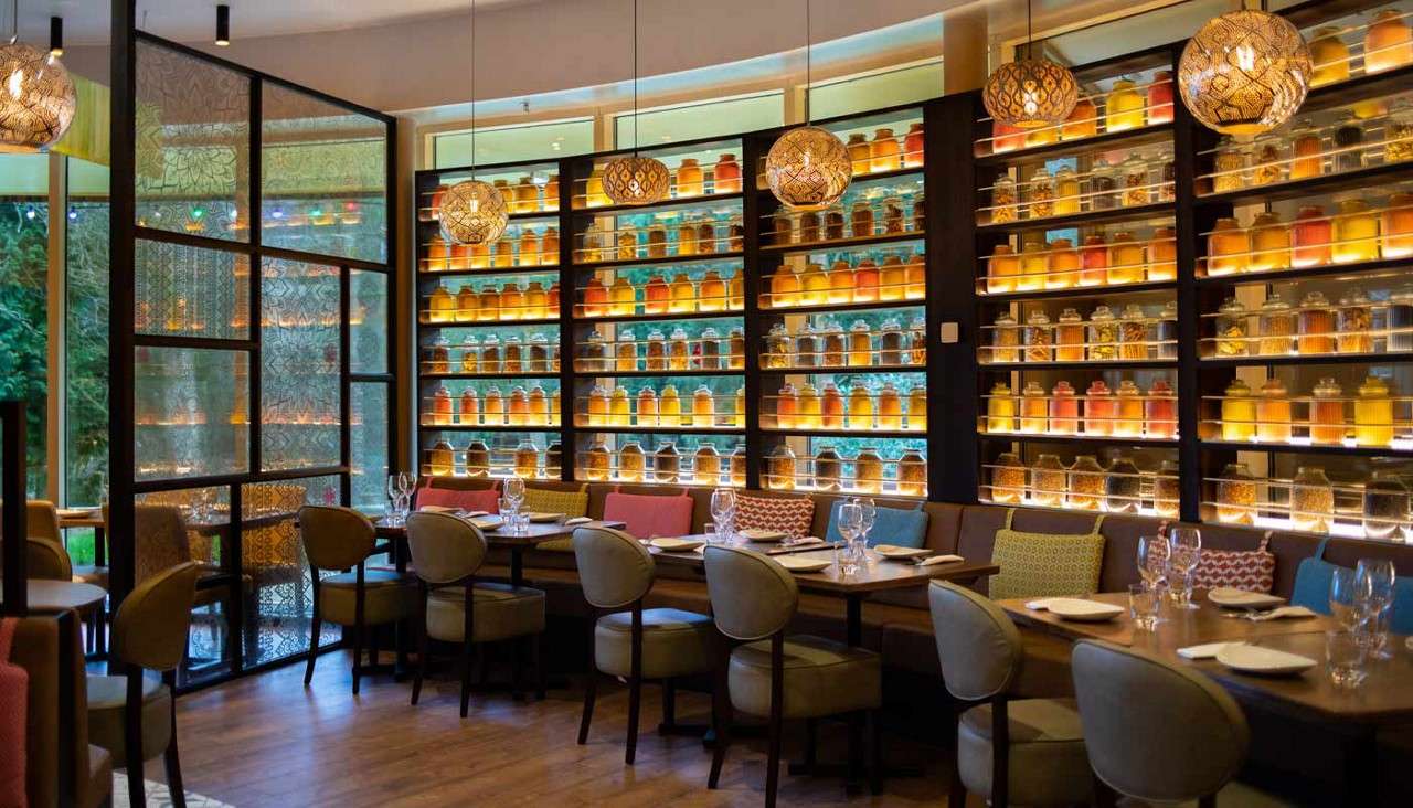 Backlit glass jars on wall shelves glow, illuminating restaurant. Tables set with plates and stemware; cushioned chairs and banquettes surround. Pendant lights hang above, while a patterned partition separates spaces.