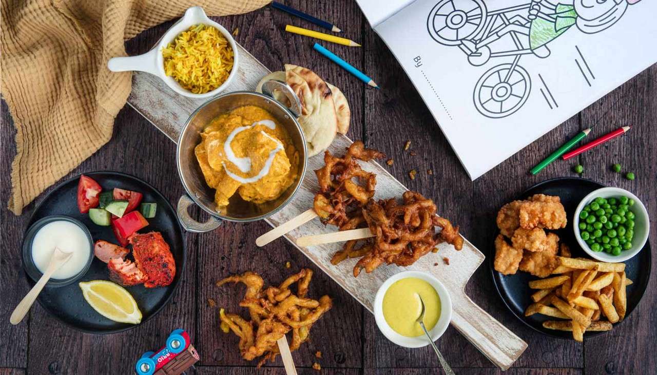 Assorted kid-friendly dishes sit spread on a wooden table—curry with naan, rice, pakoras on sticks, nuggets with fries and peas, salad, and sauces—beside crayons and a coloring book. Text: By ___________; BRIO.