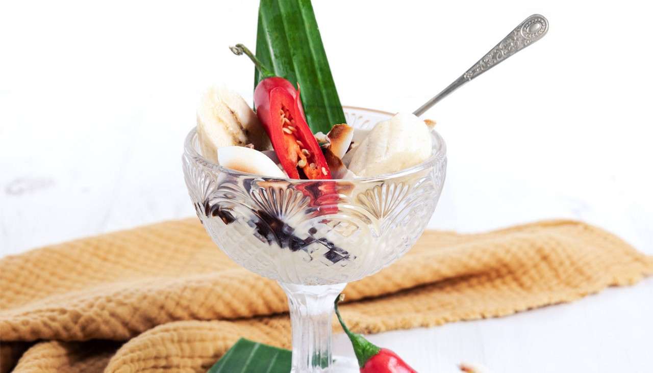 Dessert sundae in a cut-glass goblet, topped with banana slices, halved red chili, toasted coconut, and green leaf, with spoon; chocolate syrup streaks in glass; on white surface beside towel.