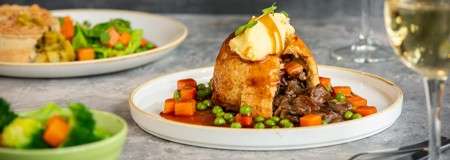 Cut-open meat pie releases beef stew, topped with mashed potatoes and gravy, surrounded by peas and diced carrots in sauce; side vegetables and a wine glass in the background.