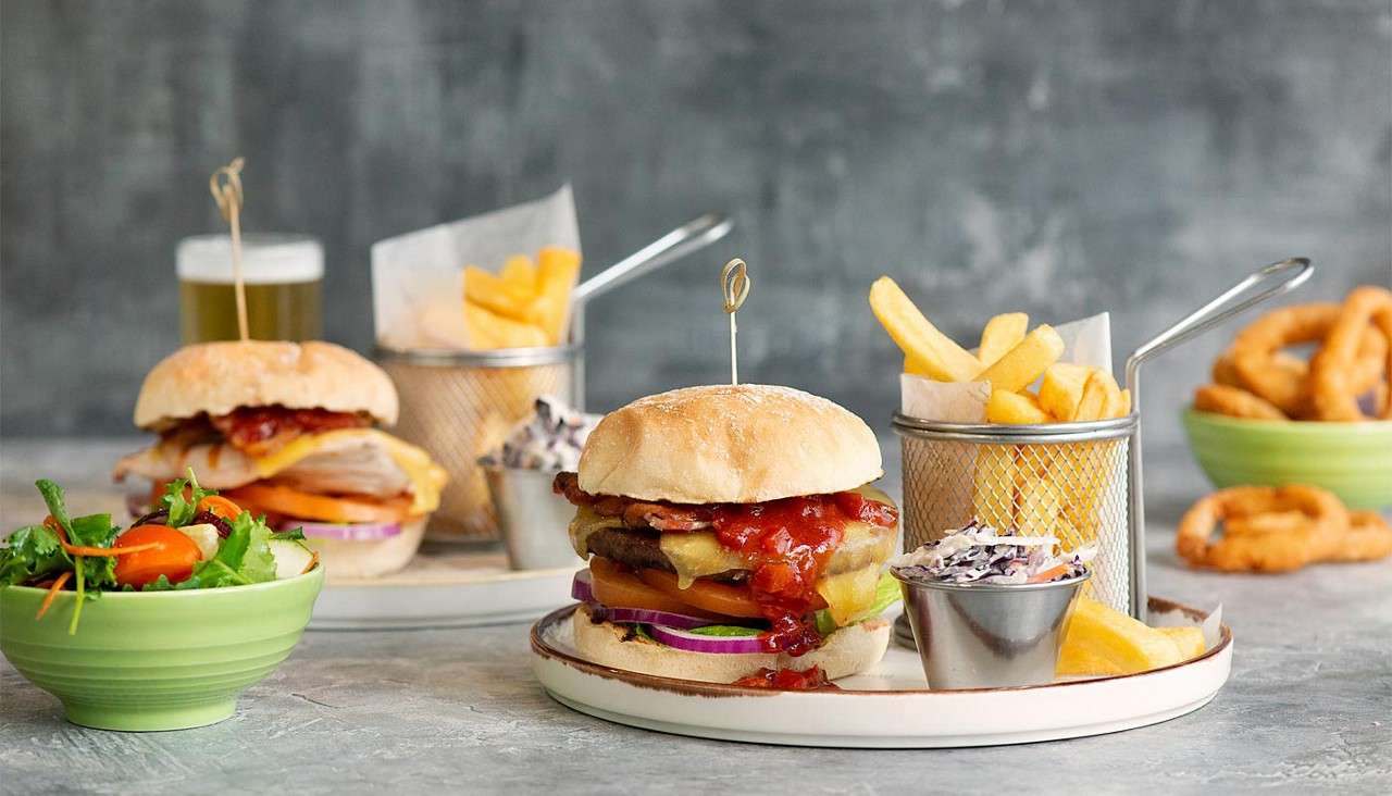Burger with cheese, bacon, and sauce drips onto a plate, accompanied by fries in a mesh basket and coleslaw; background shows burger, beer, salad, and onion rings on a tabletop.