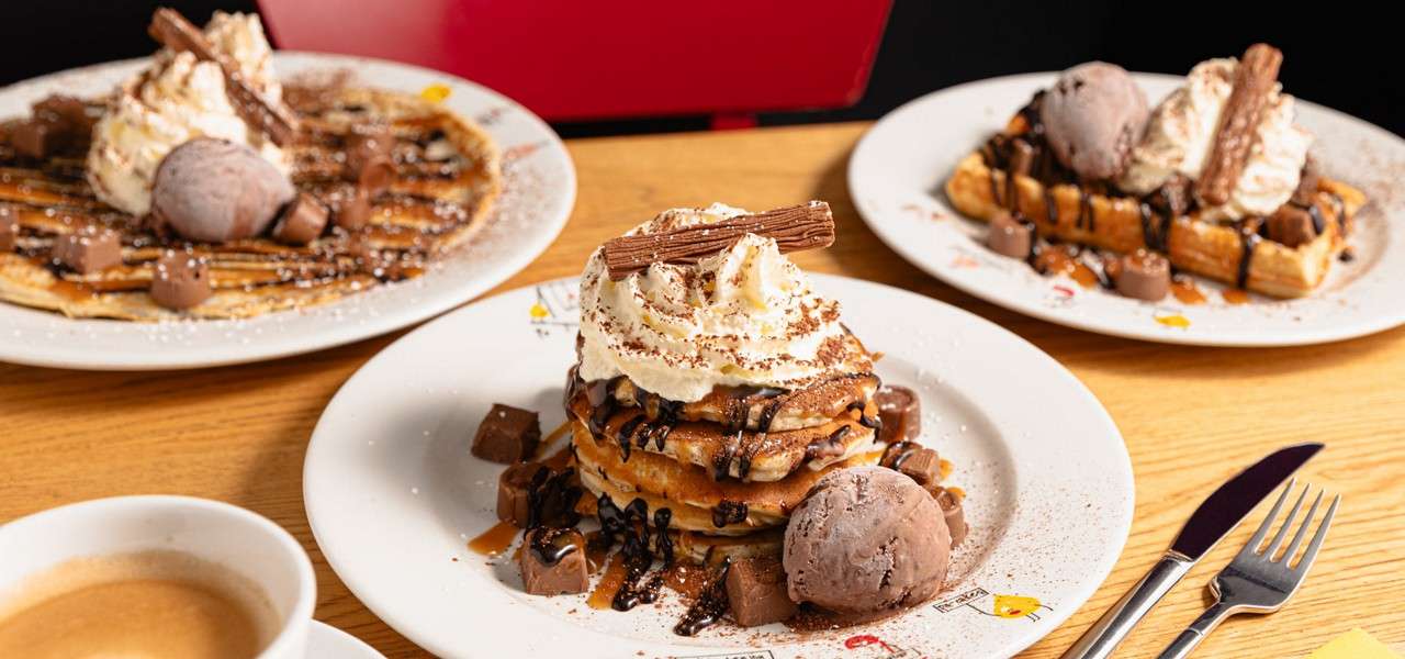 Stacked pancakes drizzled with chocolate and caramel sit on plate, topped with whipped cream and a flake bar, plus chocolate ice cream; similar waffle plates surround on wooden table.