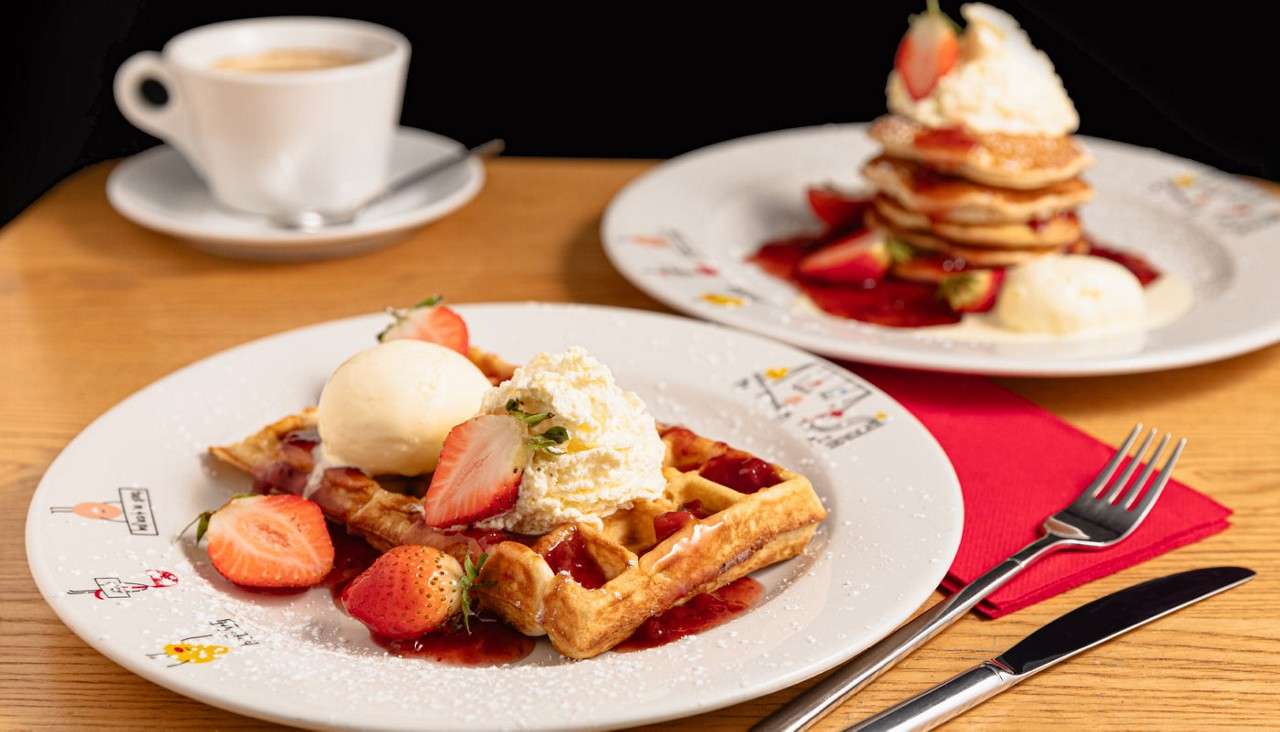 Belgian waffle with whipped cream, ice cream, and sliced strawberries sits in strawberry sauce; café setting with fork, knife, red napkin, coffee cup, and background pancake stack. Small plate doodles with unreadable text.