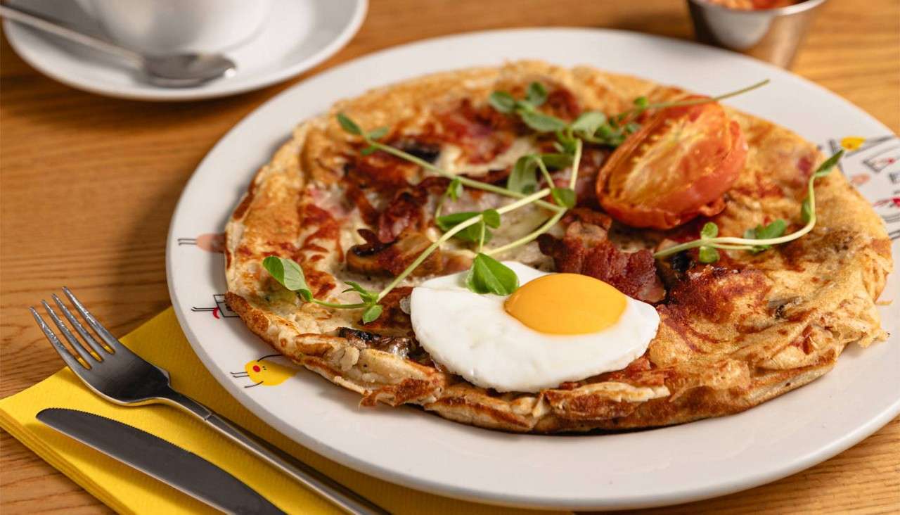 Savory pancake topped with sunny-side egg, bacon, mushrooms, tomato, and microgreens, resting on a white plate. Sits on a wooden table beside fork, knife, yellow napkin, and a coffee cup.