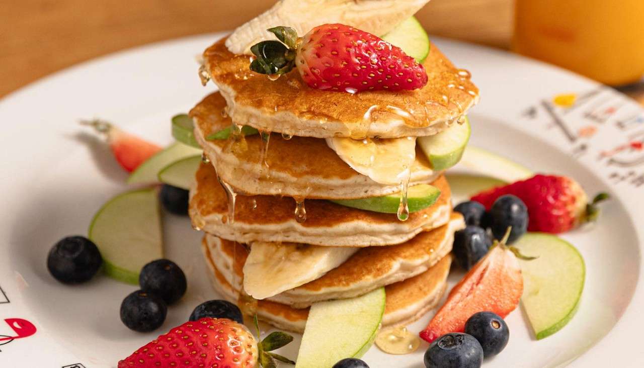 Stacked pancakes drip syrup, topped with strawberries and banana slices, surrounded by blueberries and apple wedges on a white plate, set on a wooden table with an orange drink nearby.