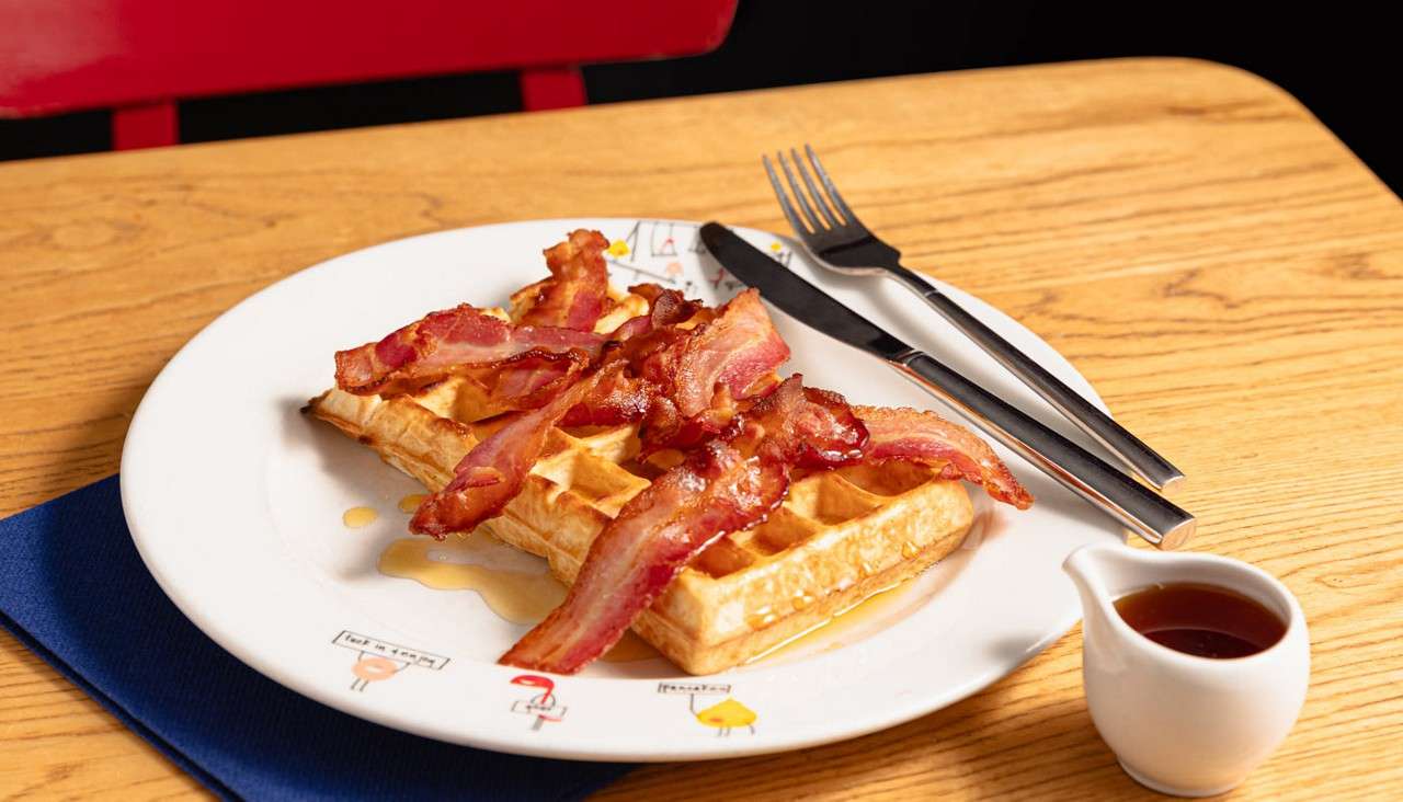 Belgian waffle topped with crispy bacon drips syrup on a white plate. A fork and knife sit beside it on a wooden table with a syrup pitcher and blue napkin.