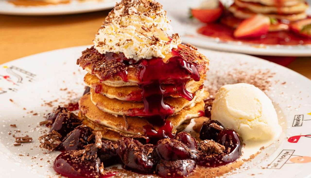 Stack of pancakes drips cherry sauce and supports whipped cream, while chocolate shavings scatter. Around it, cherries and a vanilla ice-cream scoop rest on a white plate on a table.