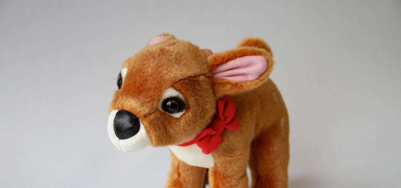 Plush fawn stands facing left, head tilted forward. Brown fur, big eyes, black nose, pink inner ears, white muzzle, faint spots, and a red bow at the neck. Light-gray backdrop.