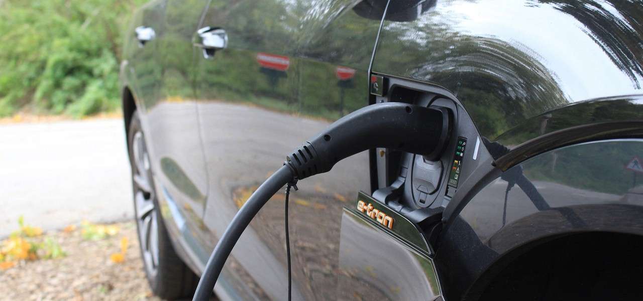 Electric car charges, a thick cable plugged into its side port labeled “e-tron,” while the vehicle sits beside a quiet, tree-lined road with scattered fallen leaves.