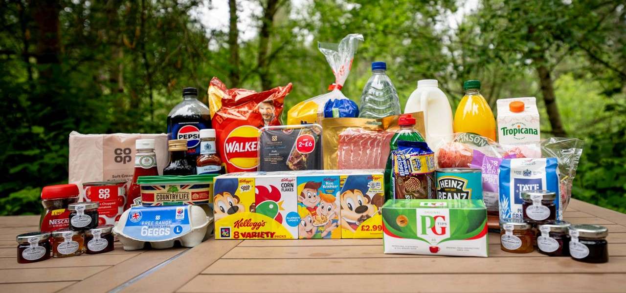Groceries arranged in rows on a wooden picnic table, displayed for viewing in a leafy forest setting.

Visible text: Pepsi; Walkers; NESCAFÉ Original; co‑op; Country Life; Free‑range Eggs L; Kellogg’s 8 Variety packs; Corn Flakes; Rice Krispies; £2.99; 2 for £6; Heinz Beanz; PG tips 40; Tropicana; TATE & LYLE PURE GRANULATED.