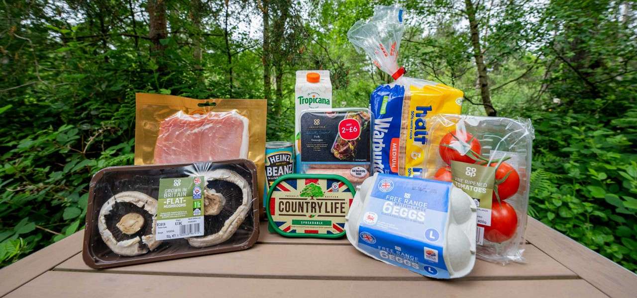 Groceries arranged upright on a picnic table; packages displayed; dense green woodland surrounds. Text: “GROWN IN BRITAIN FLAT MUSHROOMS”; “Heinz Beanz”; “COUNTRY LIFE SPREADABLE”; “Tropicana”; “ANY 2 for £6”; “Warburtons”; “half & half MEDIUM”; “co op”; “VINE RIPENED TOMATOES”; “400g”; “FREE RANGE BRITISH LARGE 6 EGGS L.”
