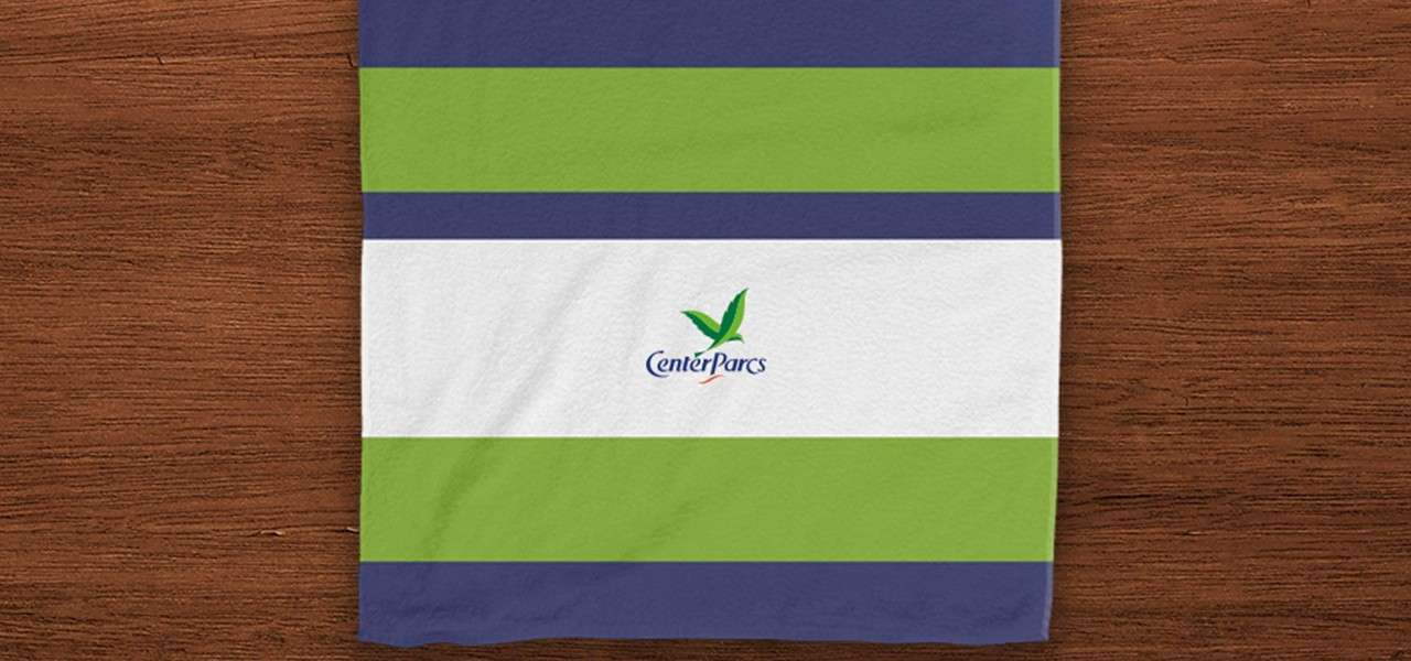 Striped towel rests flat on a wooden table; centered logo reads "CenterParcs." Horizontal bands run top-to-bottom: navy, green, navy, white with logo, green, navy.