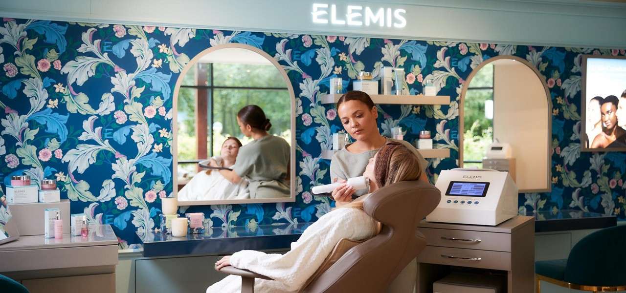 Esthetician uses a handheld device on a reclining client in a spa room with floral wallpaper, mirrors, and skincare displays. Visible text: ELEMIS, ELEMIS.