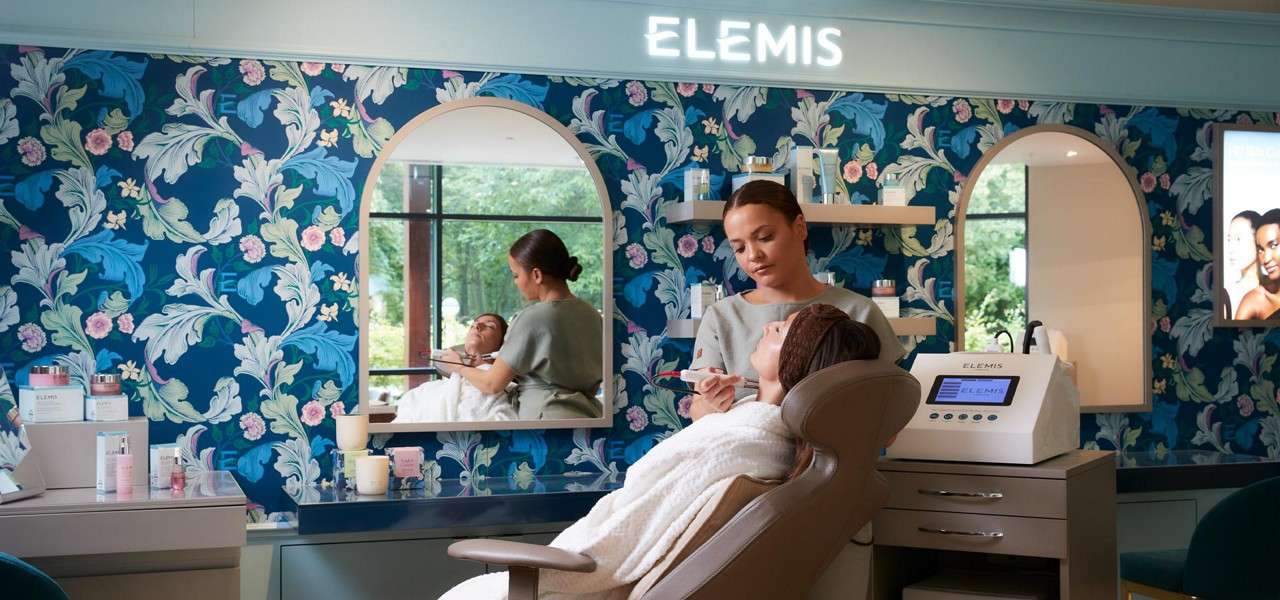 Esthetician treats a reclining client, applying product to the face, in a floral-wallpaper spa with arched mirrors, skincare shelves, and a treatment machine. Text: ELEMIS (sign, device, screen, product labels).